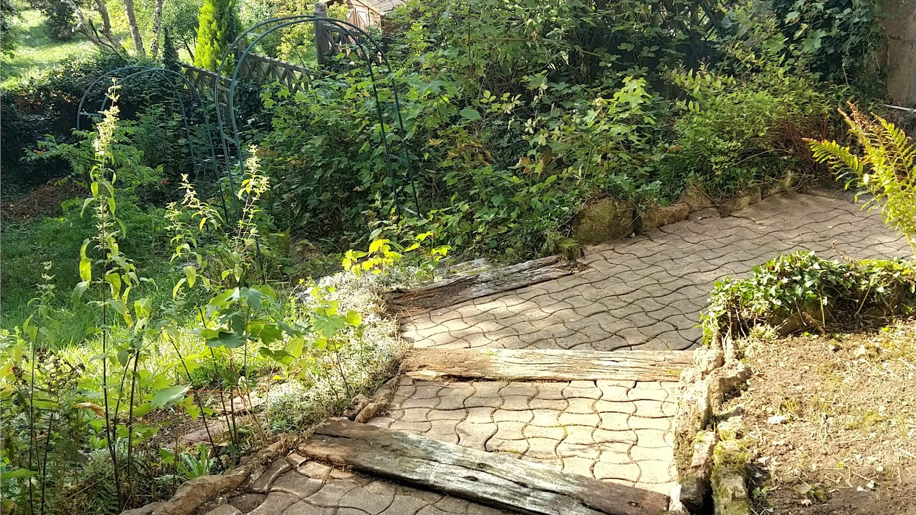 Jardin à Drouville : allée pavée, traverses en bois, arceaux et massifs au soleil
